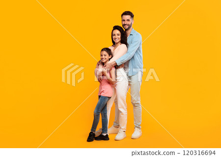 A family of three poses together against a bright yellow background. The father, mother, and young daughter are all smiling and looking at the camera 120316694