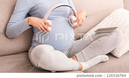 Unrecognizable Pregnant Woman Holding Headphones Near Belly Listening Music On Phone Sitting On Sofa Indoor. Cropped 120316764