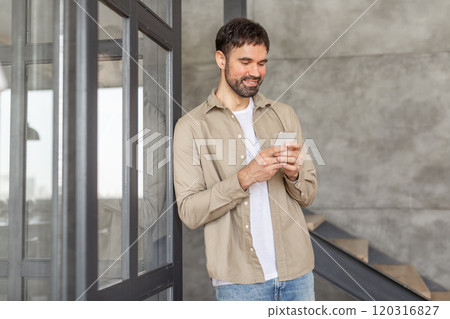 A man with a beard, wearing a tan shirt over a white t-shirt and jeans, smiles as he scrolls through his smartphone. He is standing next to a modern building with a glass door and concrete walls. A man with a beard, wearing a tan shirt over a white t-shirt and jeans, smiles as he scrolls through his smartphone. He is standing next to a modern building with a glass door and concrete walls. 120316827