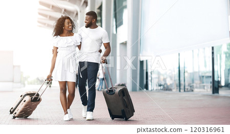 A happy African American couple, both wearing white, walks hand-in-hand through an airport terminal, pulling rolling suitcases behind them, copy space 120316961