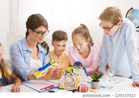 A teacher and four students are working together to build a robot in a classroom setting. All focused on the task at hand, with the teacher guiding the students and helping them with the assembly 120317015