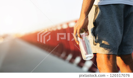 A man in athletic wear holds a water bottle in his right hand. The person is in the foreground, and the background is blurry. A man in athletic wear holds a water bottle in his right hand. The person is in the foreground, and the background is blurry. 120317076