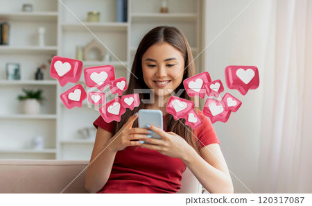 A young Korean woman in a red shirt is sitting on a couch and smiling while looking at her smartphone. Pink heart icons are surrounding her and her phone. A young Korean woman in a red shirt is sitting on a couch and smiling while looking at her smartphone. Pink heart icons are surrounding her and her phone. 120317087