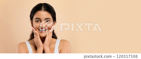 Skincare Concept. Beautiful smiling young indian woman touching face and looking at camera, attractive eastern lady enjoying her flawless skin, standing over beige studio background, copy space 120317108