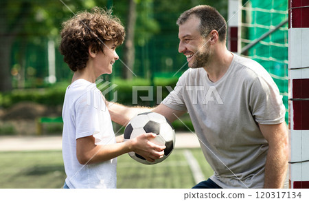 Children And Outdoor Activity Concept. Side view of smiling curly boy holding soccer ball, looking at sportive adult man who tapping and patting son on shoulder, family playing football at pitch 120317134