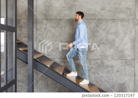 A man in a blue shirt and jeans walks up a modern interior staircase with wooden steps and black metal railings. He smiles as he ascends the stairs, which are set against a concrete wall. A man in a blue shirt and jeans walks up a modern interior staircase with wooden steps and black metal railings. He smiles as he ascends the stairs, which are set against a concrete wall. 120317197
