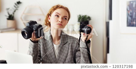 A woman in a gray blazer holds two digital cameras, considering which one to choose. 120317198