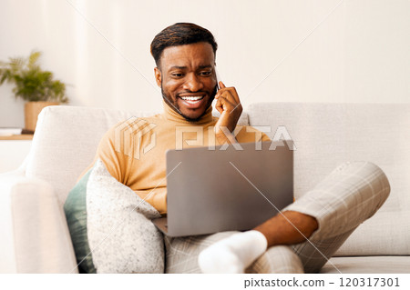 Smiling Afro Freelancer Guy Working On Laptop Computer And Chatting On Phone Sitting On Sofa At Home. Empty Space Smiling Afro Freelancer Guy Working On Laptop Computer And Chatting On Phone Sitting On Sofa At Home. Empty Space 120317301