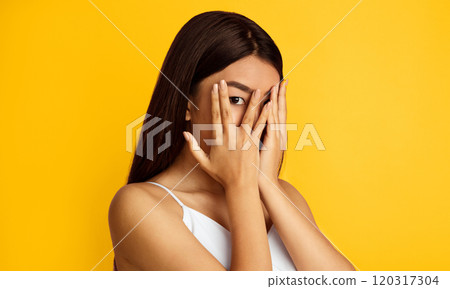 A young Asian woman with long, dark hair covers her face with her hands in a gesture of shyness or embarrassment. She is standing in front of a bright yellow background 120317304