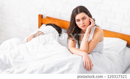 A young woman sits up in bed, looking thoughtful and sad. She rests her head on her hand, while her partner sleeps soundly beside her. The image suggests a sense of loneliness 120317321