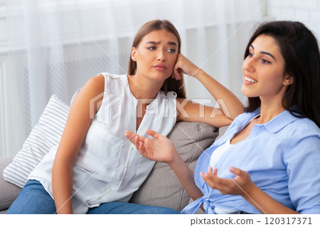 Envy. Jealous Girl Listening To Her Friend Talking About Her Great Life Sitting On Couch At Home. Selective Focus Envy. Jealous Girl Listening To Her Friend Talking About Her Great Life Sitting On Couch At Home. Selective Focus 120317371