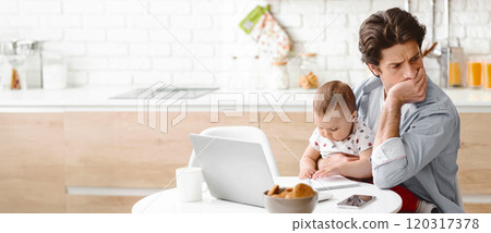 A tired father is working from home while holding his infant son. The father is sitting at a kitchen table with a laptop open in front of him, while the baby is sitting on his lap, copy space 120317378