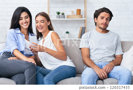 Relationship Vs Friendship. Girl Talking With A Female Friend Not Paying Attention To Her Bored Boyfriend Sitting Together On Sofa Indoor. 120317435