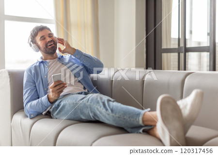A man sits on a gray sofa, wearing headphones and a blue button-down shirt, while holding a smartphone in his hand. He has his legs propped up on the sofa and is smiling while listening to music. A man sits on a gray sofa, wearing headphones and a blue button-down shirt, while holding a smartphone in his hand. He has his legs propped up on the sofa and is smiling while listening to music. 120317456