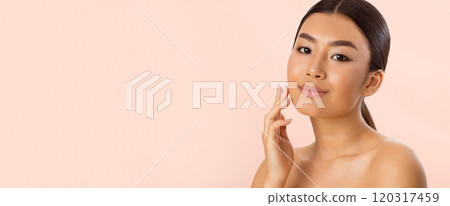 A young Asian woman with long dark hair smiles gently as she touches her face. She is standing against a light pink background. Her skin appears smooth and healthy, copy space A young Asian woman with long dark hair smiles gently as she touches her face. She is standing against a light pink background. Her skin appears smooth and healthy, copy space 120317459