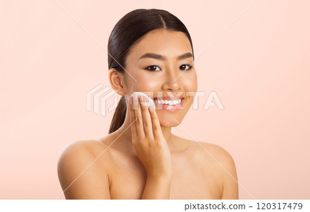 This image shows a young Asian woman with long dark hair smiling as she applies makeup to her face with a cotton pad. The background is a light pink color. This image shows a young Asian woman with long dark hair smiling as she applies makeup to her face with a cotton pad. The background is a light pink color. 120317479