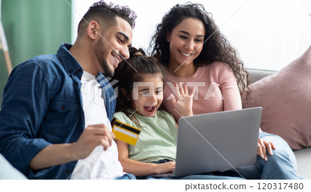 Cheerful Arabic Family Of Three With Laptop And Credit Card Making Online Shopping While Relaxing On Couch At Home, Happy Middle Eastern Parents And Little Daughter Enjoying Purchase From Internet Cheerful Arabic Family Of Three With Laptop And Credit Card Making Online Shopping While Relaxing On Couch At Home, Happy Middle Eastern Parents And Little Daughter Enjoying Purchase From Internet 120317480