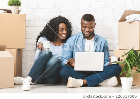 Moving, relocating concept. Positive african couple purchasing furniture for new apartment, sitting on floor and using laptop, panorama with free space 120317500