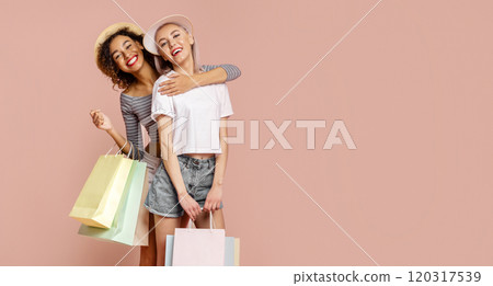This image features two young women, one Black and one White, who are smiling and hugging each other. They are both dressed in casual summer clothing and are holding shopping bags. 120317539