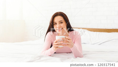 A woman is laying on a bed, holding a cup in her hands. The soft sheets and pillows provide a comfortable backdrop as she enjoys a moment of relaxation with her beverage. 120317541