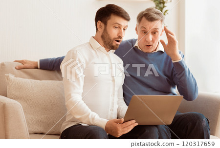 Oops, computer problem. Shocked elderly man and his adult son using laptop looking at error message sitting on sofa indoor Oops, computer problem. Shocked elderly man and his adult son using laptop looking at error message sitting on sofa indoor 120317659