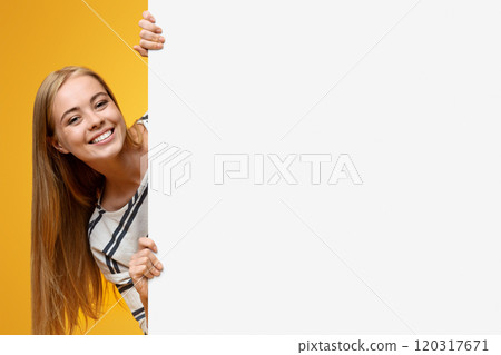 Cute teenage girl looking out of white blank placard for your text or product, yellow background 120317671