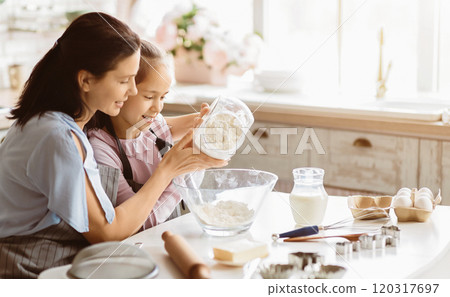 A mother and her young daughter are in a kitchen, working together to bake. The daughter is pouring flour from a glass container into a bowl, while her mother watches and smiles. A mother and her young daughter are in a kitchen, working together to bake. The daughter is pouring flour from a glass container into a bowl, while her mother watches and smiles. 120317697