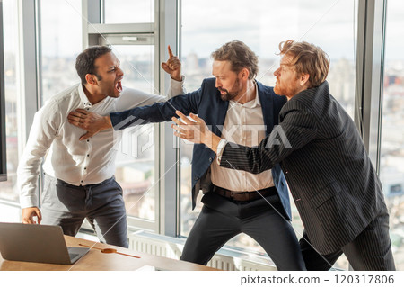 Three men in suits are engaged in a heated argument in a modern office setting. The man in the white shirt is yelling at the man in the blue suit, who is pushing him away 120317806
