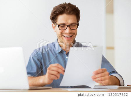 A man wearing glasses smiles and laughs while reading a document at his desk. 120317822