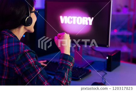 A gamer celebrates their victory in a dimly lit room with neon lighting. They have headphones on and are pumping their fist in the air. The word victory is displayed brightly on computer monitor. A gamer celebrates their victory in a dimly lit room with neon lighting. They have headphones on and are pumping their fist in the air. The word victory is displayed brightly on computer monitor. 120317823