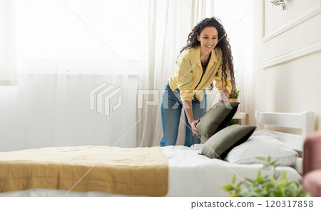 Cheerful young millennial lady making bed at home, holding pillow, doing housework in the morning. Positive stylish woman cleaning up her bedroom, everyday routine concept, free copy space 120317858
