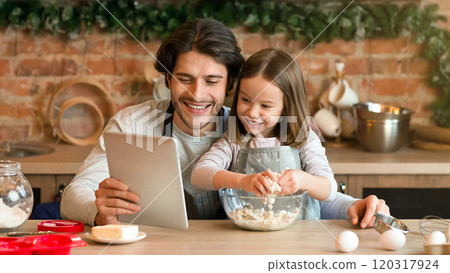 Happy dad and little daughter checking recipe of dough online on digital tablet while cooking pastry in kitchen together, free space Happy dad and little daughter checking recipe of dough online on digital tablet while cooking pastry in kitchen together, free space 120317924