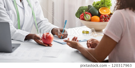 Dietician african woman giving young patient healthy diet tips, holding fresh red apple, clinic interior Dietician african woman giving young patient healthy diet tips, holding fresh red apple, clinic interior 120317963
