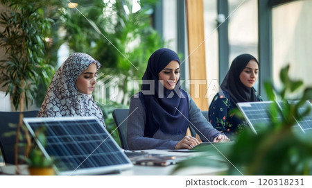 Three women wearing headscarves sit at a table working. Two of the women are facing the camera, with one looking at her tablet and the other looking away Three women wearing headscarves sit at a table working. Two of the women are facing the camera, with one looking at her tablet and the other looking away 120318231