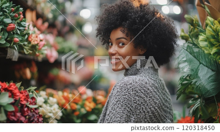 A joyful woman with curly hair looks back with a smile while exploring a lively flower market filled with blooming plants and vibrant colors during daylight. The atmosphere is cheerful and inviting. 120318450