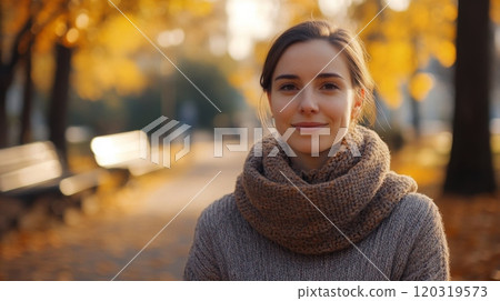 A young woman stands confidently in a park adorned with vibrant fall colors. She wears a warm sweater and a thick scarf, surrounded by golden leaves and soft sunlight filtering through trees. A young woman stands confidently in a park adorned with vibrant fall colors. She wears a warm sweater and a thick scarf, surrounded by golden leaves and soft sunlight filtering through trees. 120319573