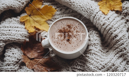 A steaming mug of hot chocolate sits atop a knitted blanket, accompanied by colorful autumn leaves, creating a warm, inviting atmosphere. 120320717