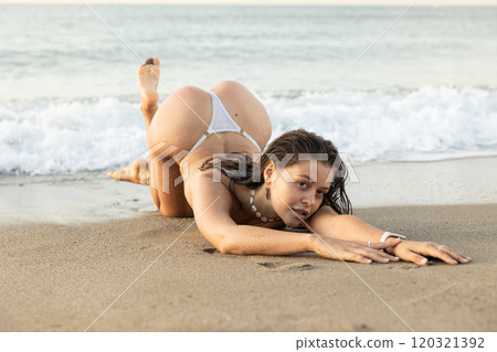 Young woman in thong posing while kneeling on beach Young woman in thong posing while kneeling on beach 120321392