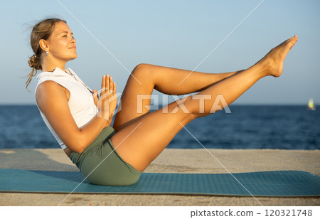 On windy day young slender woman does yoga near sea doing Ubhaya Padangushthasana 120321748