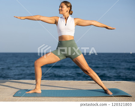 Sportive girl practicing warrior pose of yoga on seashore Sportive girl practicing warrior pose of yoga on seashore 120321803
