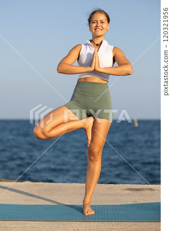 On windy day young slender woman does yoga near sea doing Vrikshasana 120321956