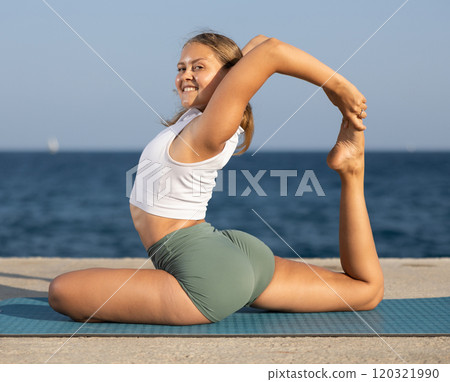 On windy day young slender woman does yoga near sea doing royal pigeon pose 120321990