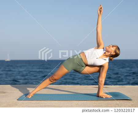 On windy day young slender woman does yoga near sea doing Parivritta Parshvakonasana 120322017