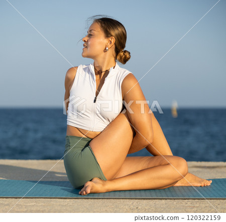 Flexible girl practicing lord of the fishes pose of yoga on seashore Flexible girl practicing lord of the fishes pose of yoga on seashore 120322159