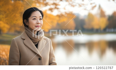 A woman stands calmly by a lake, surrounded by vibrant autumn leaves. She is dressed warmly and appears contemplative, appreciating the beauty of the changing season in a serene park setting. 120322787