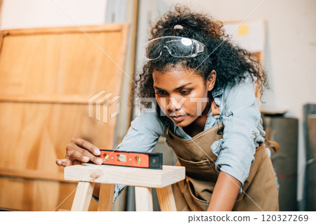 African American carpenter woman uses a level on a wooden stool in the workshop. Master joiner skillfully crafting solid pine furniture, surrounded by tools and materials, National Carpenters Day 120327269