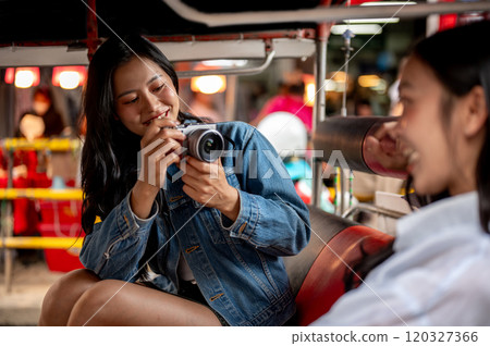A happy Asian girl takes a picture of her friend with a camera while enjoying a tuk-tuk ride. A happy Asian girl takes a picture of her friend with a camera while enjoying a tuk-tuk ride. 120327366