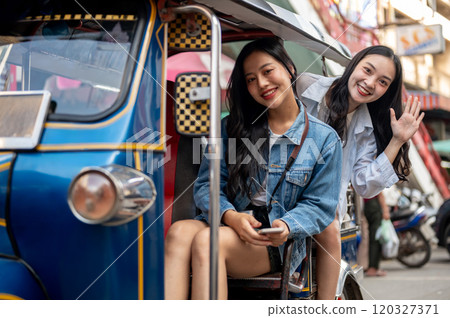 Two cheerful Asian women enjoy a tuk-tuk ride in Thailand, excited by the stunning city views. 120327371