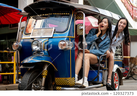 Two excited Asian girls enjoy a ride in a tuk-tuk in Thailand, amazed by the stunning views. Two excited Asian girls enjoy a ride in a tuk-tuk in Thailand, amazed by the stunning views. 120327373