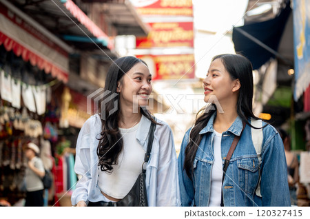 Two happy Asian female tourists explore a local market in Thailand, walking along the street. 120327415